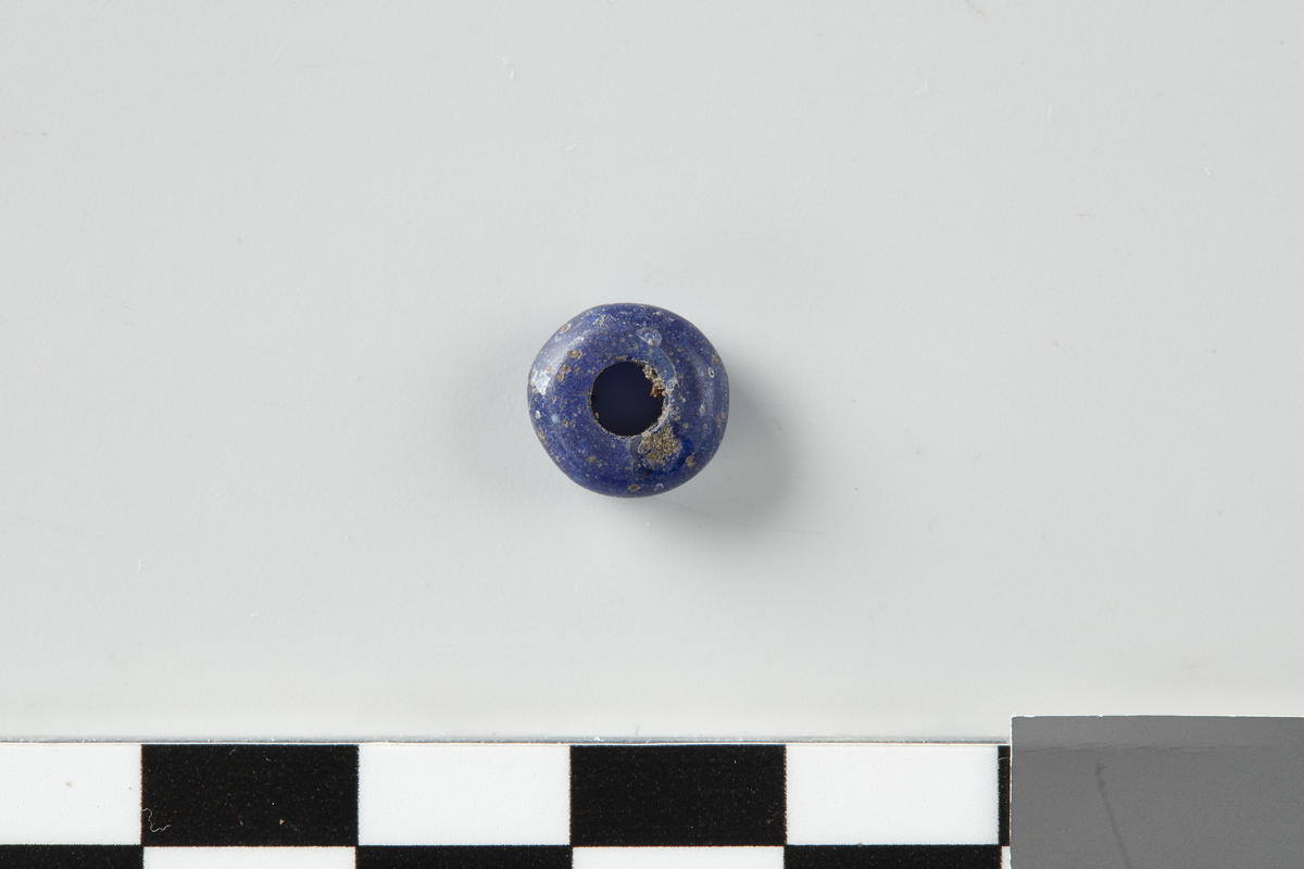 Glassperle. Rund skiveformet perle av blåfarvet glass. Br: 0,9 cm, h: 0,3 cm.