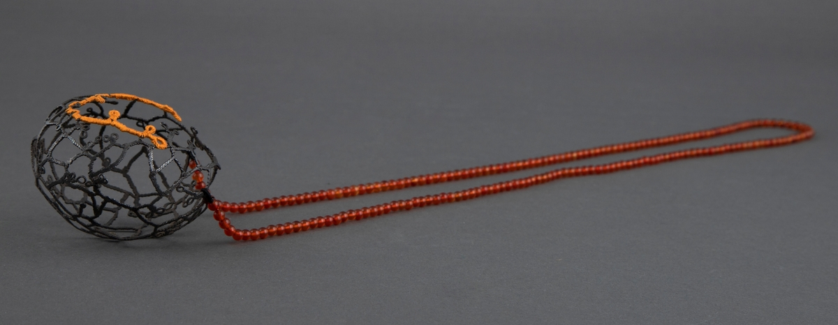 Halssmykke bestående av et kjede av små, røde runde stener og et kuleformet, gjennombrutt anheng i oksydert sølv. På fremsiden har anhenget en noe større åpning der kanten delvis er omheklet med oransje silke.