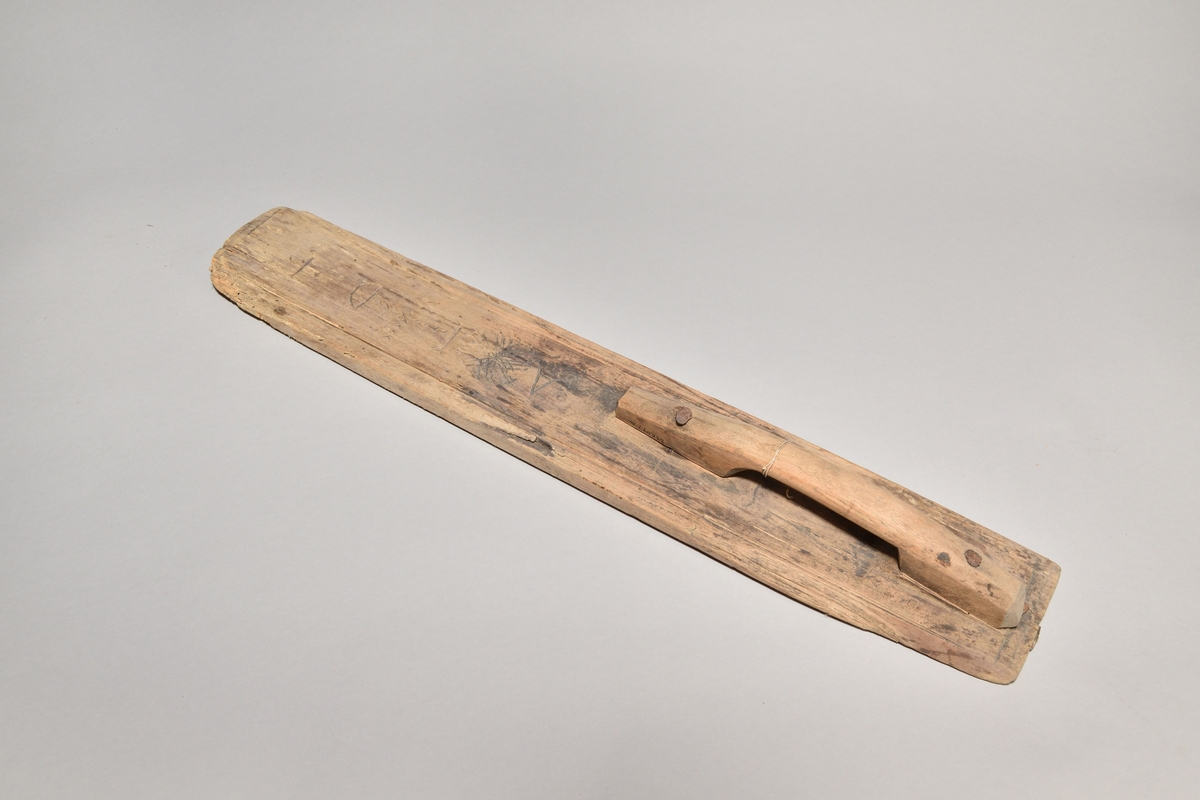 Kaveldon tillverkat i trä, med platt bräde och skuret handtag. Ovansidan av brädet med inskurna mönster. 