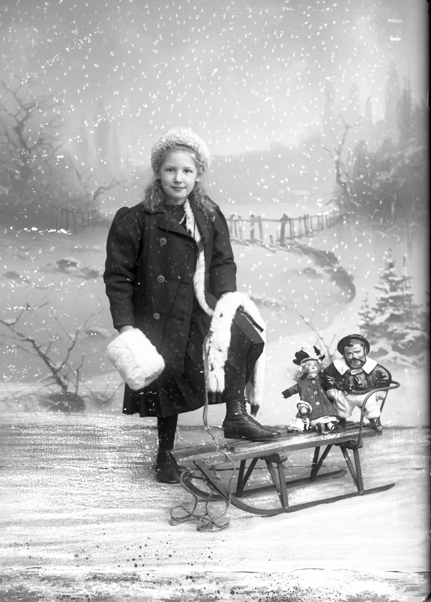 Ateljébild av fotografens systerdotter. Hon är vinterklädd mot snöfond och har ena foten på en kälke, där det sitter leksaker baktill.