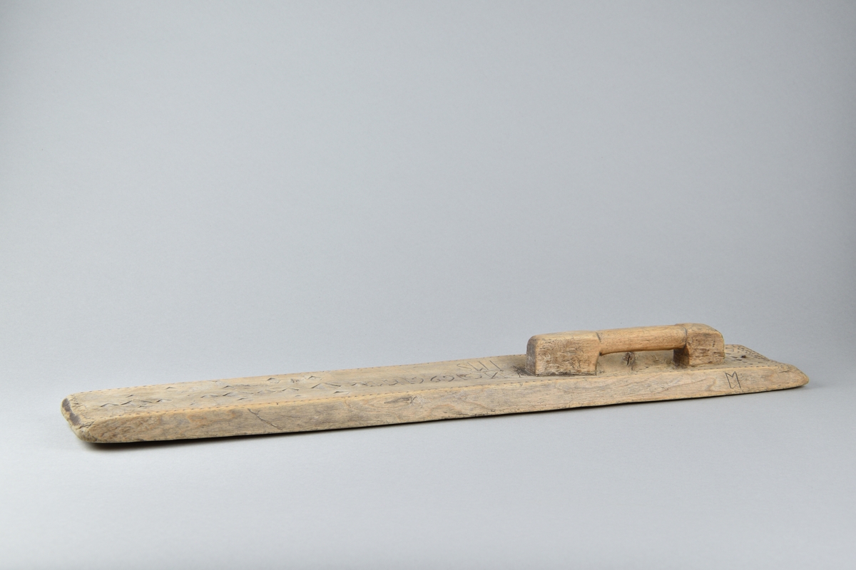 Kaveldon tillverkat i trä. Långt och fyrsidigt bräde med handtag. Ovansidan ornerad med romber och sicksack mönster, samt inskriften "ANO 1696 HKMDIMS".