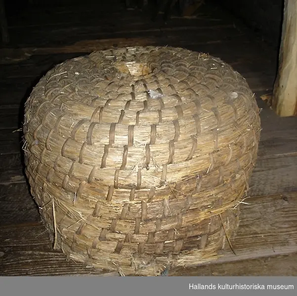 Rund bikupa av halm, flätad, med snidade delar av trä (ingång för bin). Rester av bivax på insidan.