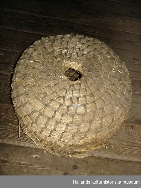 Rund bikupa av halm, flätad, med snidade delar av trä (ingång för bin). Rester av bivax på insidan.
