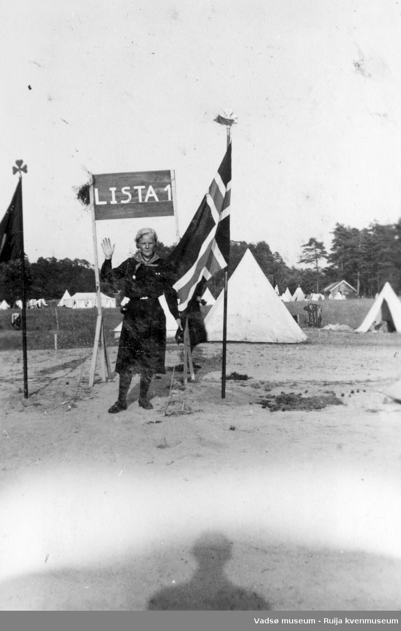 Speiderjente vinker mot fotograf. Oppstilt sammen med banner og to flagg, et av dem norsk. I bakgrunnen ses hvite telt og leirområde.