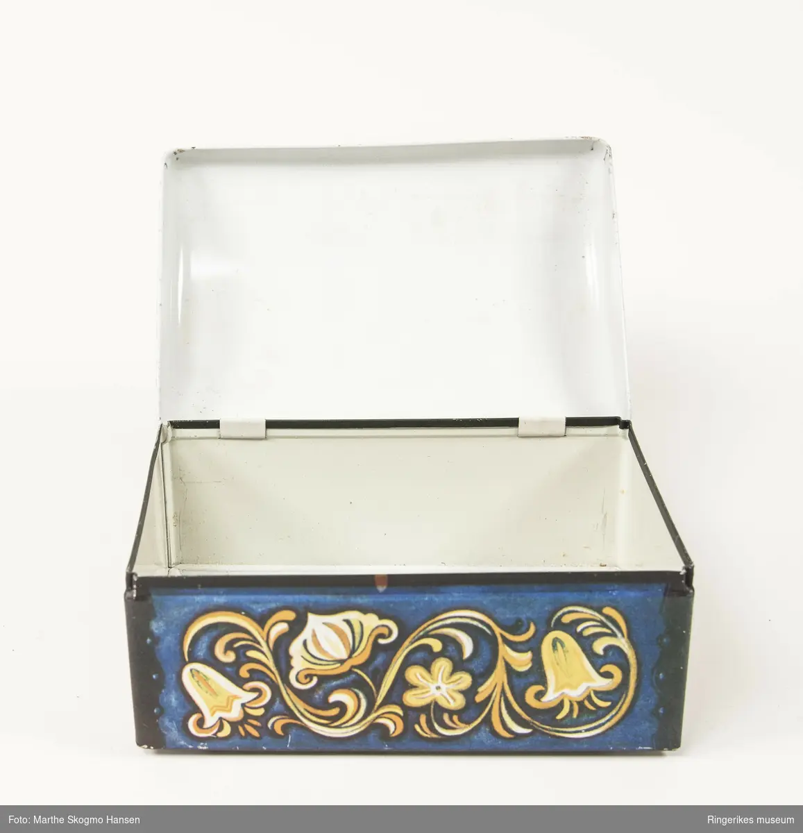 Matboks i blikk formet som en kiste med buet lokk hengslet i bakkant og dekorert for å illustrere en kiste med rosemaling og hjørnebeslag. Under boksen er logoen til Sætre kjeks i uthevet relieff.