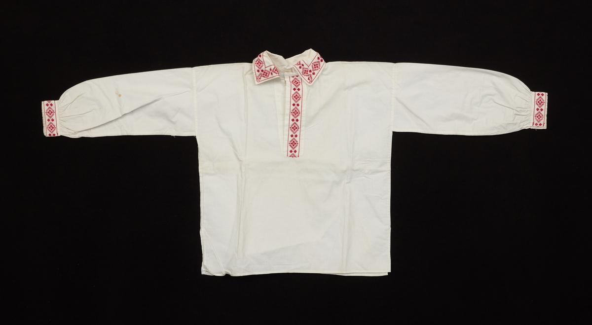 Skjorta till komponerad sockendräkt för barn. Av vitt bomullstyg. Sprund, krage och ärmlinningar är dekorerade med mönstervävt röd/vitt band. Knäpps med metallhake och handsydda hyskor.