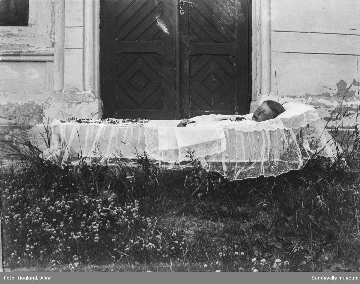 En ung avliden pojke i öppen kista framför ingången till ett hus.