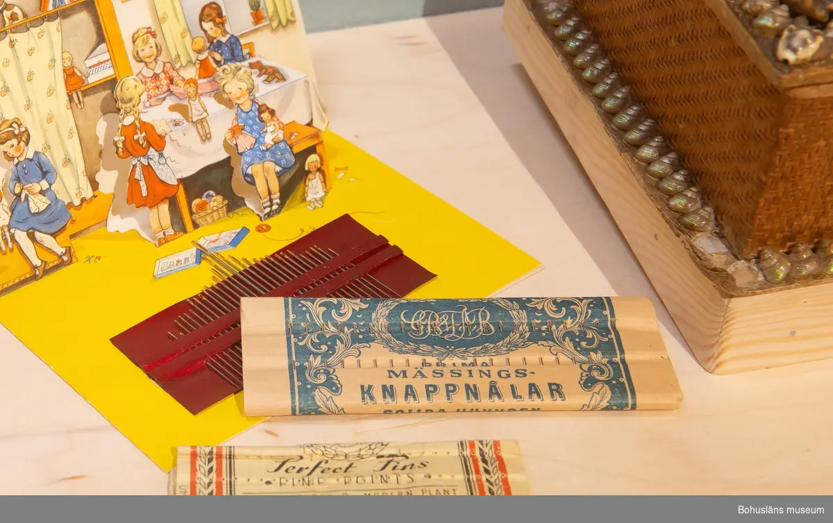 Pappersförpackning med knappnålar och text och dekor i blått tryck. Fem rader om vardera 20 knappnålar.
Text:
"Prima mässingsknappnålar. Solida  huvuden. 
Varning. I handeln förekommer utländska knappnålar av järn förvillande lika våra av mässing. Sådana nålar rosta
och förstöra det föremål var i de fästas. Köp därför aldrig knappnålar som ej bära vårt nedanstående skyddsmärke".