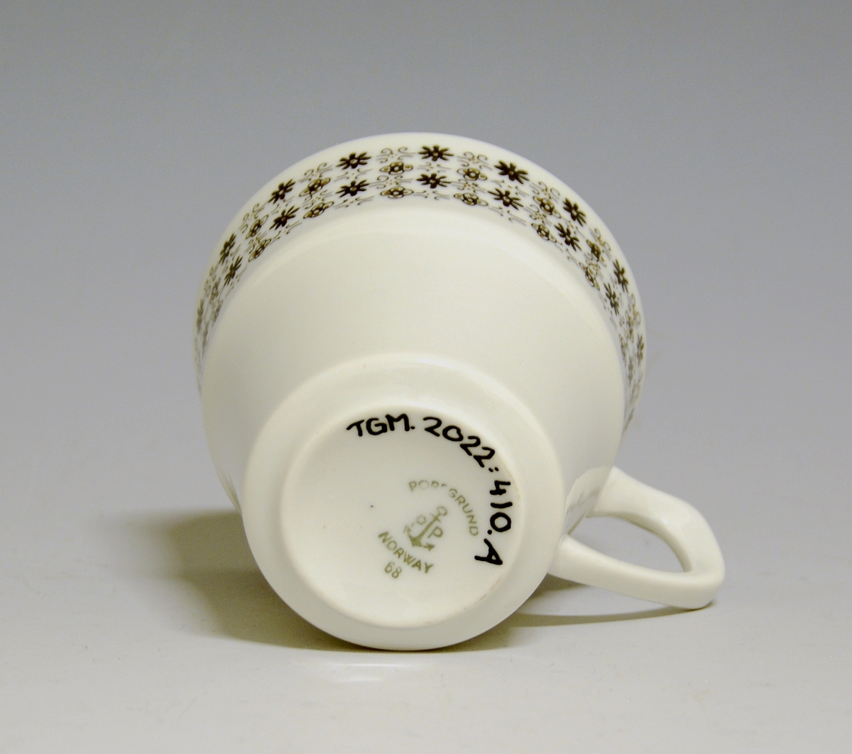 Kaffekopp av porselen med hvit glasur og bord med småblomstret trykkdekor i sort og gull i øvre del av korpus.
Modell: Petita (eller variant av)
