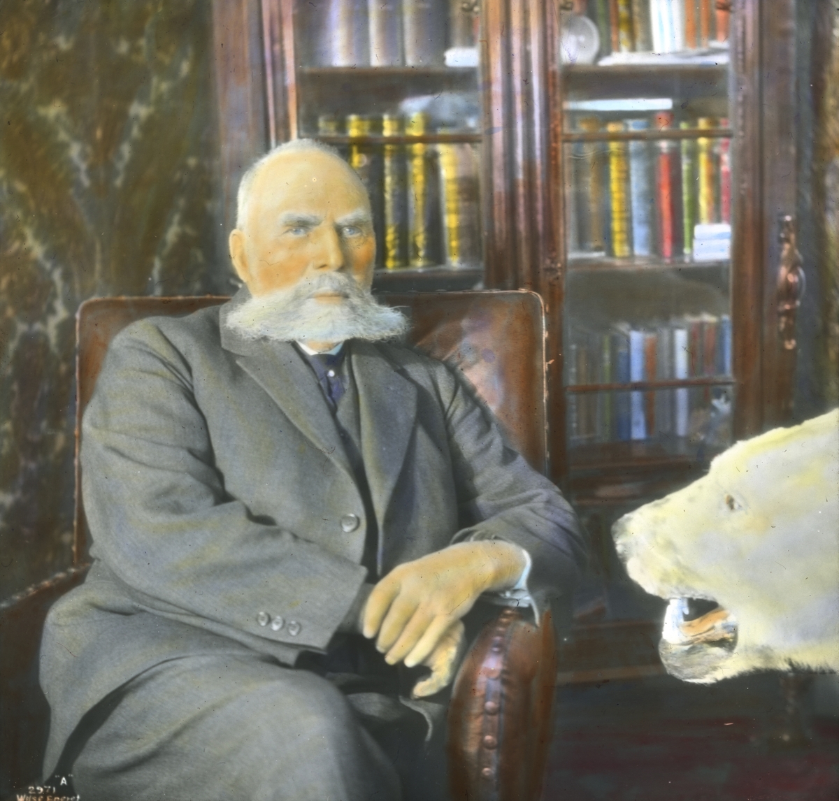 Håndkolorert dias. Bildet viser polfareren Otto Sverdrup sittende i en lenestol. I forgrunnen vises hodet til en utstoppet isbjørn.
