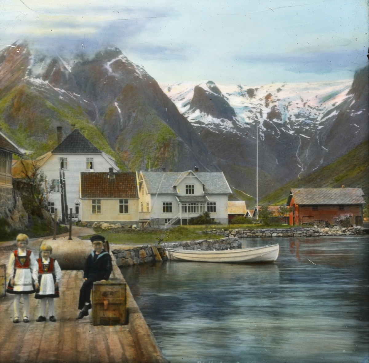 Håndkolorert dias. Bildet viser tre barn på en brygge i Balestrand. I bakgrunnen ses en robåt, trehus, grønne fjellsider og snøkledde topper. Bildets tittel "Balholm" er det tidligere navnet til tettstedet Balestrand.