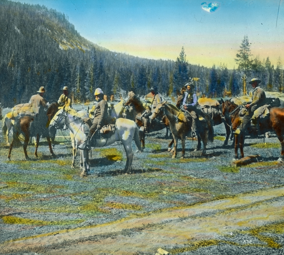 Håndkolorert dias. Åtte menn til hest i et skogs- og fjellandskap i Montana, U.S.A.