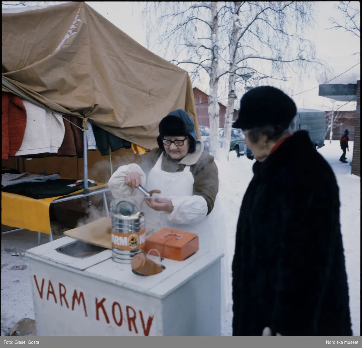 Varm korv. Korv-Inga Eriksson serverar en kund varm korv från sitt stånd på marknaden i Jokkmokk.