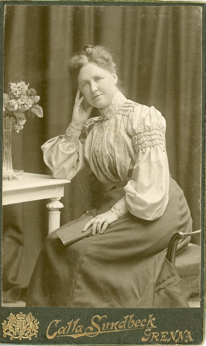 Kabinettsfotografi av en okänd kvinna som sitter vid ett bord med blommor på. Hon vilar huvudet i höger hand.