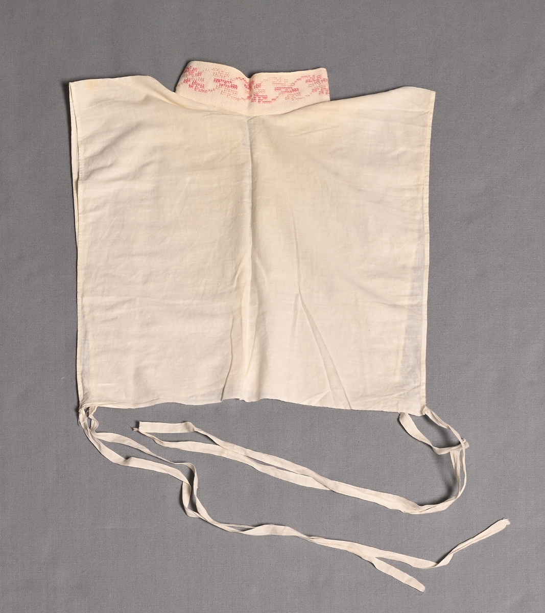 Isättning till grönländsk festdräkt för kvinnor.
Löst skjortbröst av vitt bomullstyg med liten ståkrage med rosa-blekrosa korsstygnsbroderi på aidaväv. Knyts med bomullsband i båda sidorna.