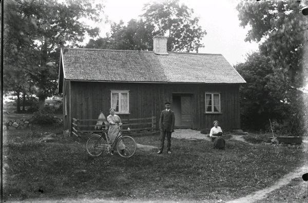 Fader Anders Johansson med döttrarna Berta och Jenny i Tränshult i trädgården. En av döttrarna håller i en cykel och den andra sitter på huk med en hund i knät. Vid stugan står en instängslad bikupa. Halva sticktaket har nyligen lagts om.