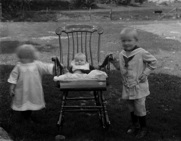 Troligen tre syskon utomhus med en bebis i gungstol mellan en pojke i randig kolt med sjömanskrage och kortbyxor och en yngre flicka.