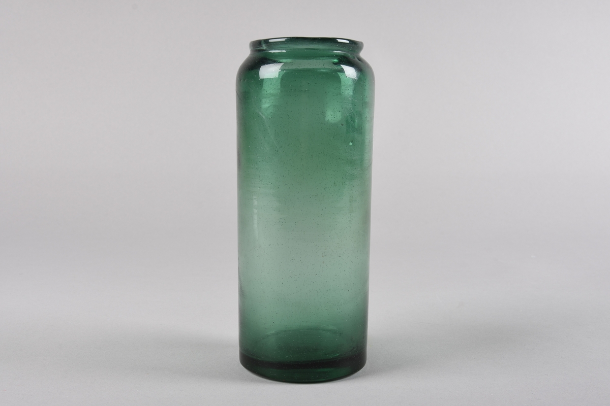 Sylindrisk støypt glaskrukke. Korpus snevrast inn i toppen og deretter ut til ein smal ujamnt forma krage ved munningsranda. Produsert av resirkulert glas. Glaset er grønfarga.
