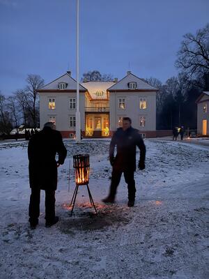 To personer (anstatte ved museet) varmer seg ved en bålpanne i parken. Snø på bakken og Eidsvollsbygningen i bakgrunnen.