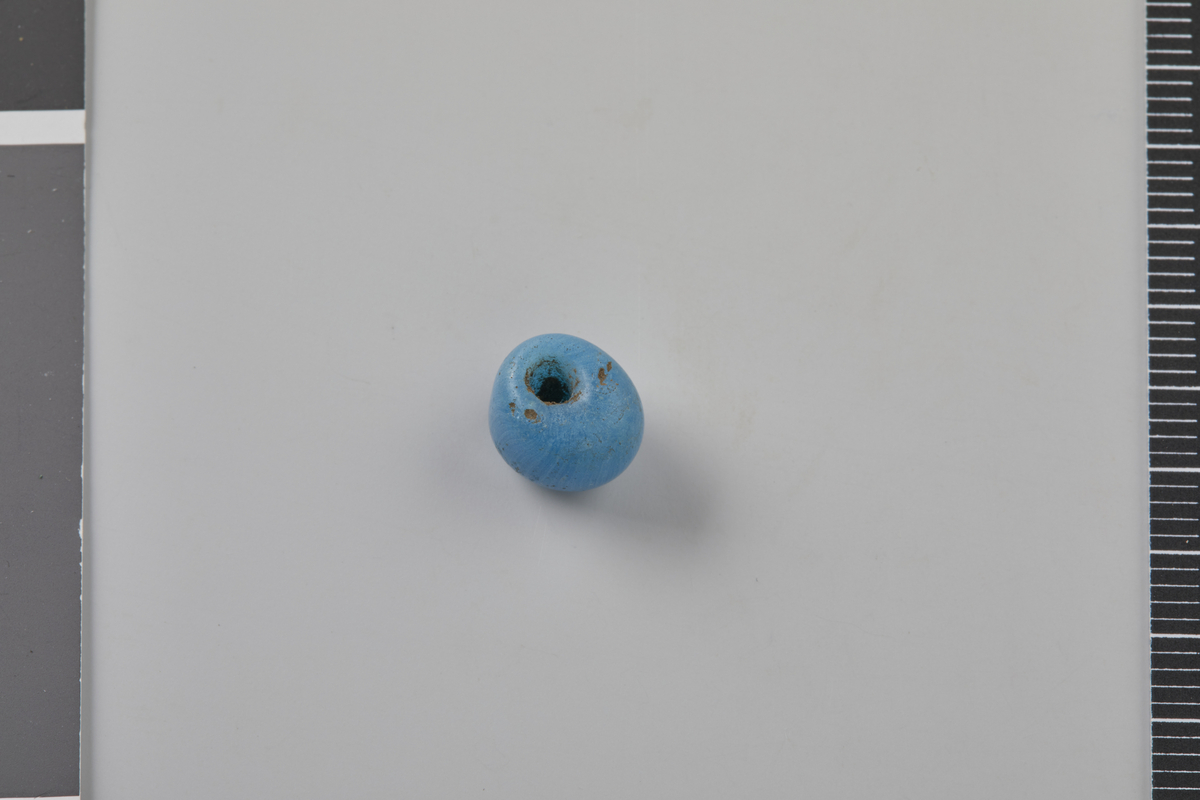 Tønneformet perle av turkist opakt glass. Lignende perler er kjent fra yngre jernalder, men kan også være yngre (jf. Vinsrygg 1979:Pl. 1-2; Thunmark-Nylén 1998:Pl. 309-310). Diam. 0,8 cm, hullets diam. 0,22 cm, h. 0,8 cm. Vekt: 1,0 g.