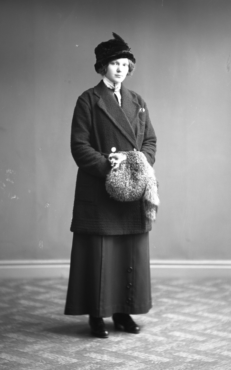 Ateljébild av ung kvinna stående i helfigur iförd ytterkläder, hatt och muff. Beställare: Anna Johansson. Fototid (osäker) 1910.