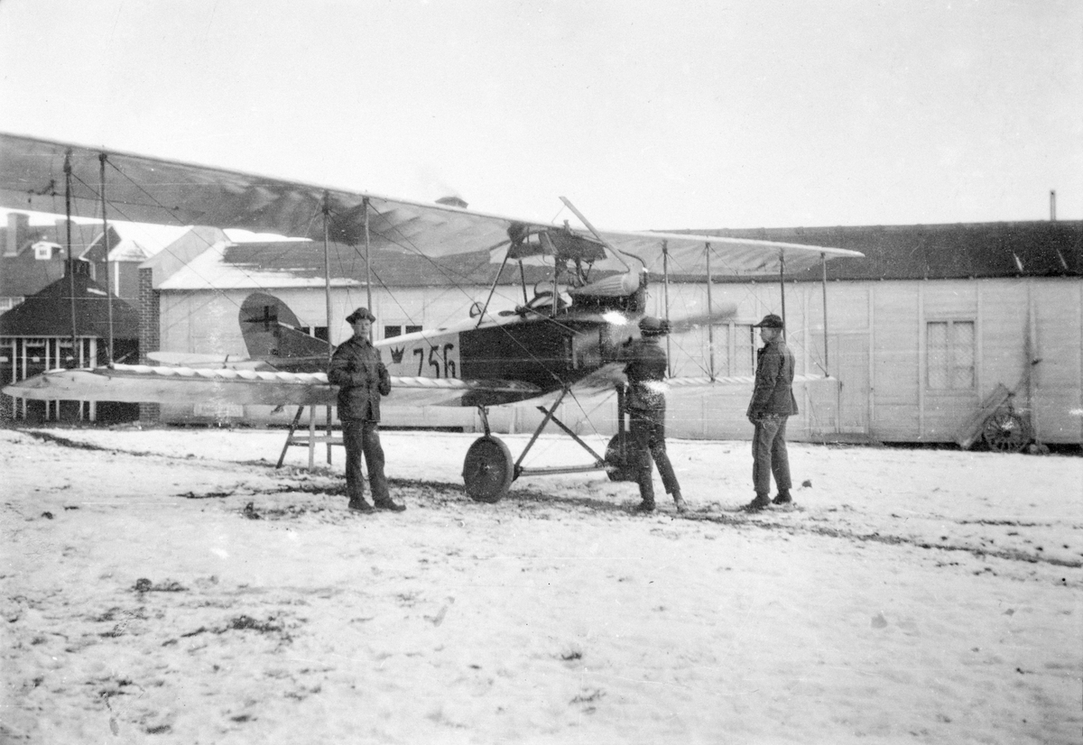 Flygplan Albatros NAB 9 nummer 756 tillhörande Flygkompaniet står uppställd på bock på flygverkstäderna Malmslätt, vintertid omkring 1917-1924. Tre militärer vid flygplanet.