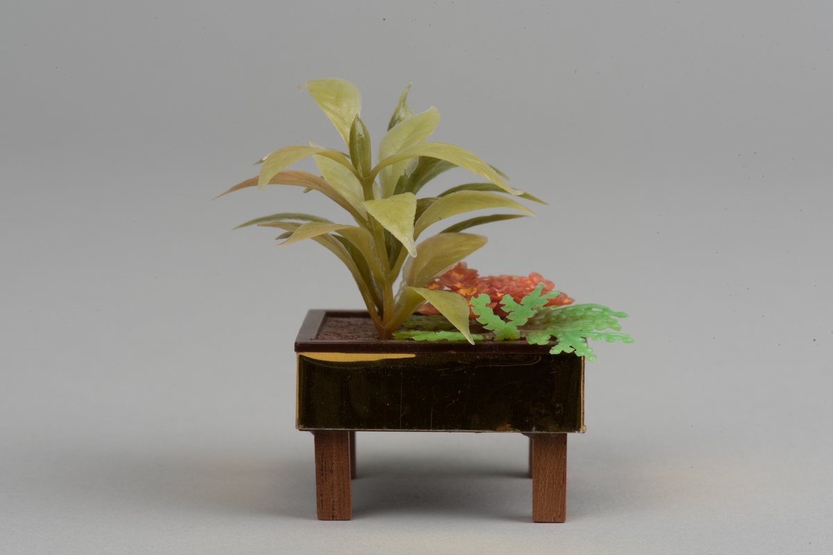 Kvadratiskt blomsterbord med gröna växter, tillverkade i plast.
Bordet har fyra ben av trä, bruna. Sidorna är guldfärgade. 
Inuti bordet står tre olika växter i gröna nyanser och en växt i bruna nyanser.
Bordet är tillverkat av Lundby, som dockskåpstillbehör.