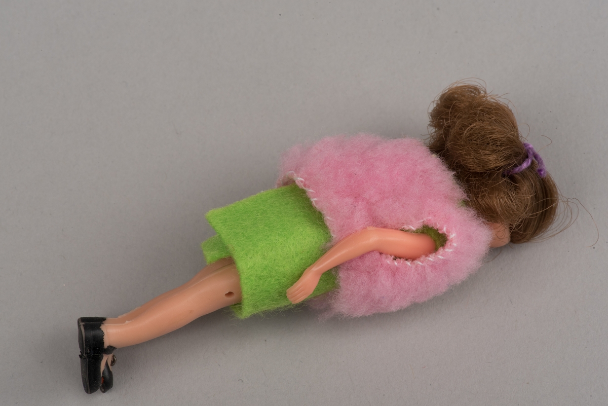 Flickdocka för dockskåp, troligen tillverkad av Lundby leksaksfabrik, i plast.
Dockan är klädd i pålimmad ärmlös klänning av grön filt och en rosa ärmlös väst. På fötterna ditmålade svarta skor.
Håret är ihophållet av en lila tråd i tofs.