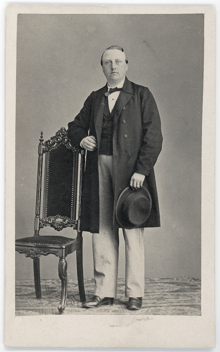 Kabinettsfotografi - en man står vid en stol med en hatt i handen, Uppsala