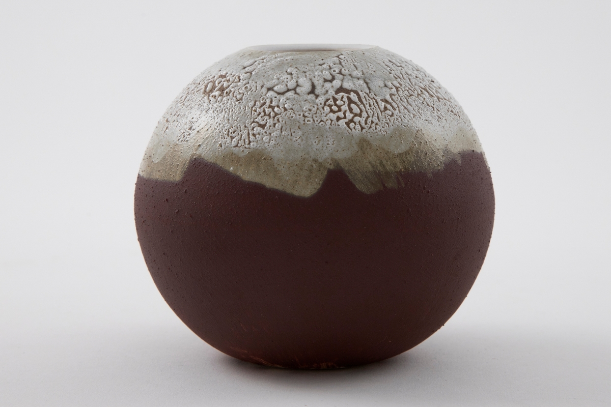 Liten rund vase i matt brun terrakotta. Tykk hvit glasur med små hull og risser på vasens øvre tredjedel. Delvis lukket form med sirkelrund munning på toppen.