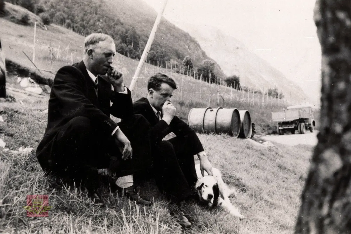 Vegmester Olav Midtkandal (til venstre) og sjåfør på vedlikehaldet Thorleif Thorsnes tar seg ein røyk, iført finkleda i vegskråninga ned mot Thorsnesgarden. Fat med drivstoff ligg på vegkanten og den franske Forden til Vegvesenet som Thorleif fekk i 1946 står borti vegen. 