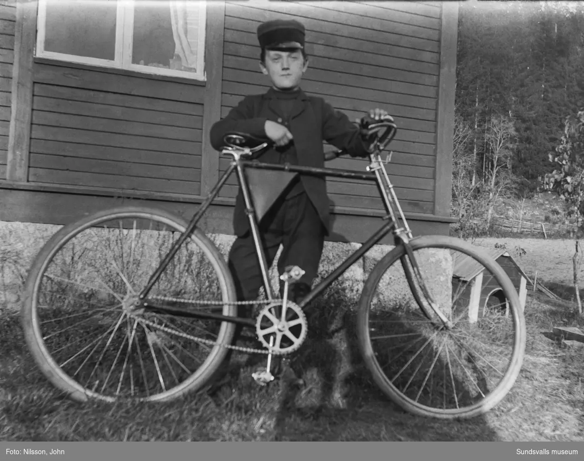 En pojke i skärmmössa står med en cykel vid husknuten till Roken 1:3 i Laggarberg.. Hundkoja i bakgrunden.