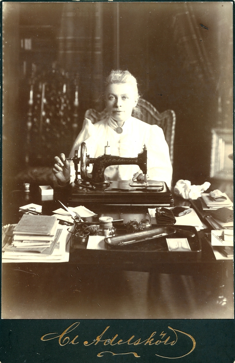 Porträtt av Gustafva Adelsköld (f. Brolin) (1844-1940) vid en symaskin och ett bord med böcker, sytillbehör mm. Fotograferad av hennes man Claes Adelsköld.