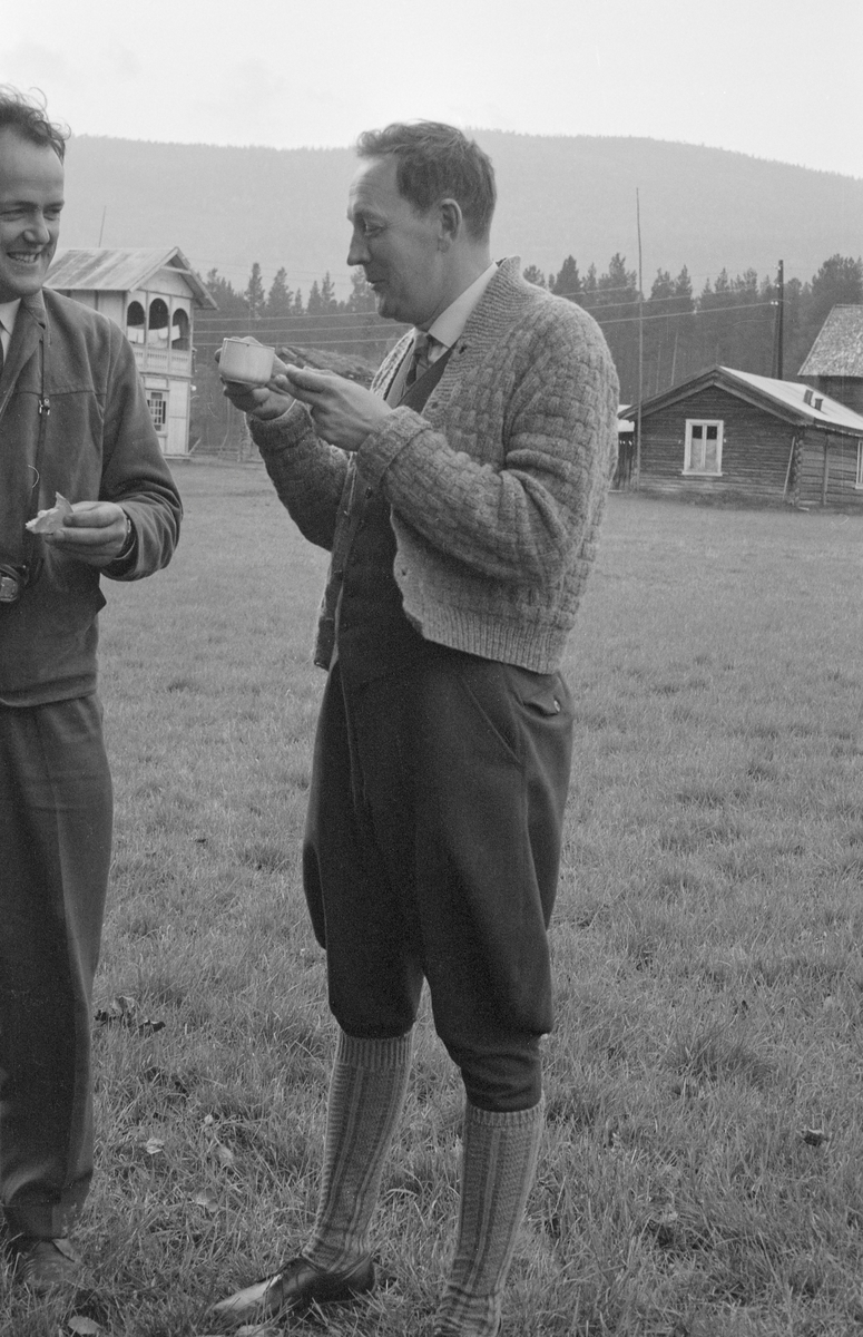 To fløtingsfunksjonærer, fotografert under befaring, antakelig ved Trysilvassdraget. Fotografiet er tatt i utkanten av ei eng, der de stående inntok et lunsjmåltid. Fotografen som deltok i befaringa har notert seg deltakernavnene Modin, Sørebø, Svensson, Akre og Svensson. Vi vet foreløpig ikke hvem mannen som står sentralt i dette bildet, men han som befinner seg ved vestre bildekant er gjenkjent som Per Ivar Akre (1925-1997). Han var andre generasjons fløtersjef i den norske delen av vassdraget. I bakgrunnen skimter vi et gardstun.