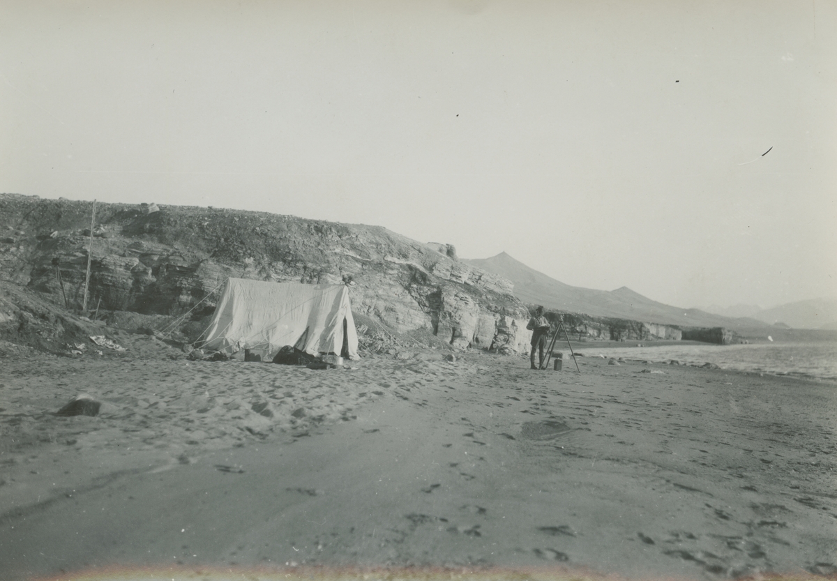 Fotografi från expedition till Grönland. Motiv av en man, troligen Otto Nordenskjöld, som står på en karg strand med berg i bakgrunden. Till vänster om honom står ett vitt tält uppställt.