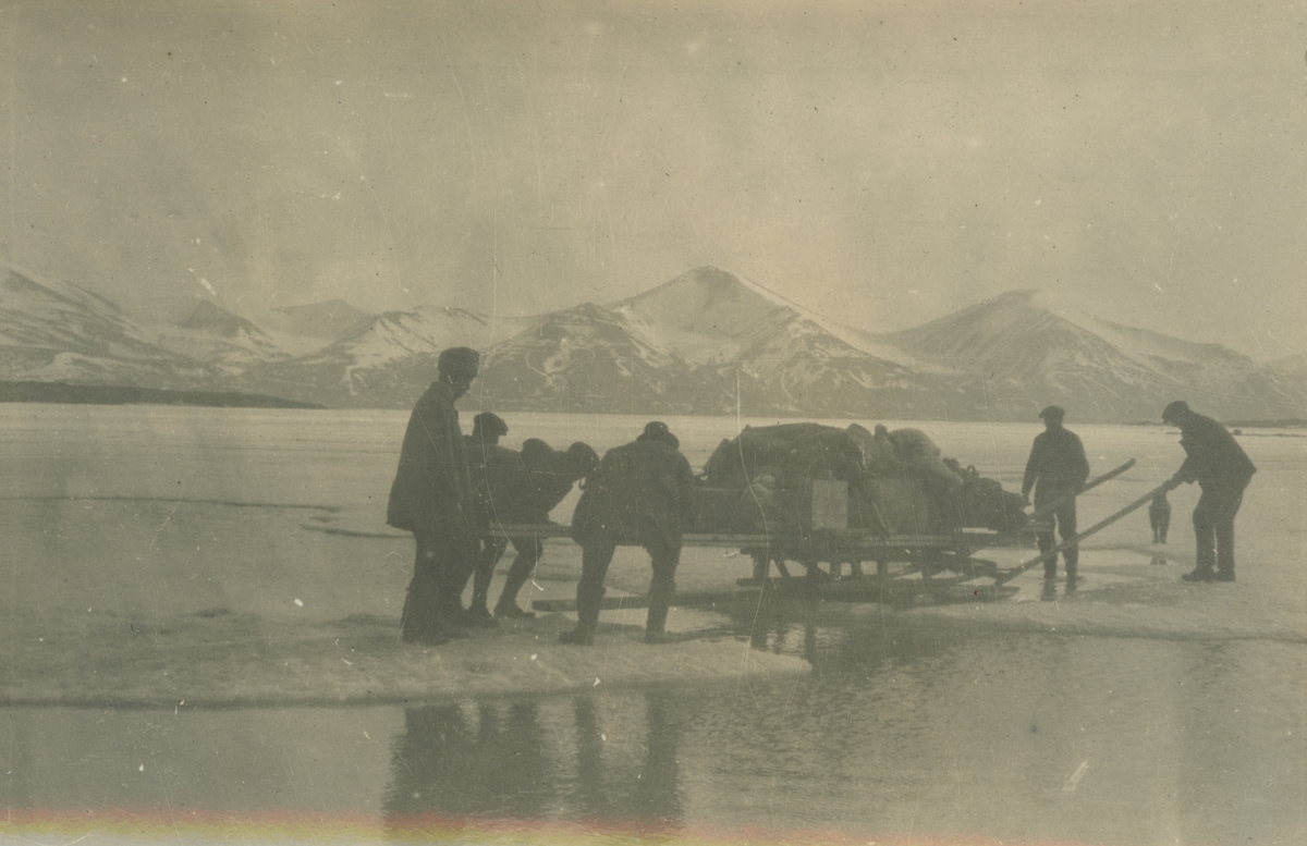 Fotografi från expedition till Spetsbergen. Motiv av ett antal expeditionsdeltagare som drar en fullastad släde över isen. I bakgrunden syns vidsträckta snötäckta berg.