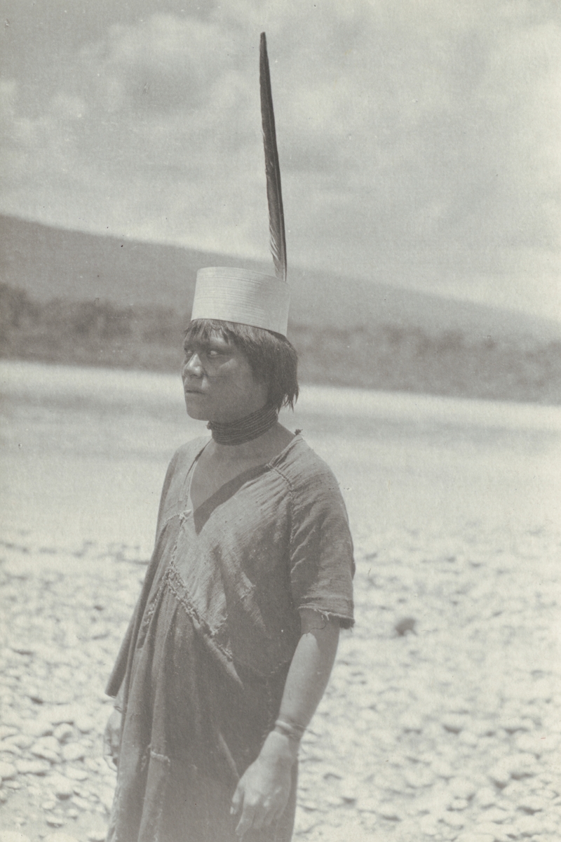 Fotografi från kuvert märkt med "Ernst Nordenskjöld". Motiv av man iklädd hatt med fjäder vid flod i djungel.