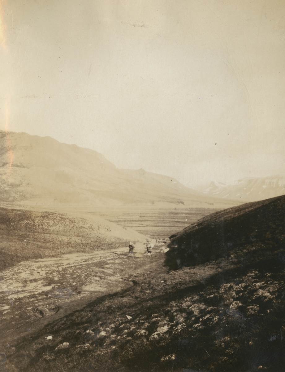 Fotografi från expedition till Sveagruvan. Vy över ett kargt och vidsträckt bergslandskap. I mitten av bilden syns två fullpackade expeditionsdeltagare som vandrar.