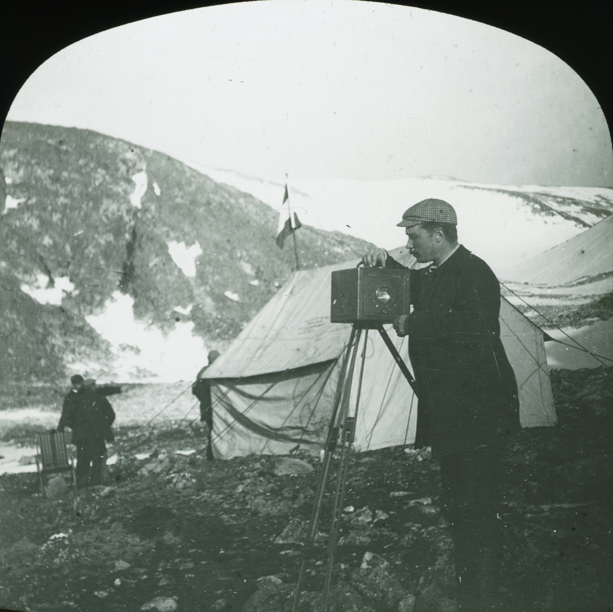 Glasnegativ med motiv av Nils Strindberg som står och ställer in sin kamera. I bakgrunden skymtas ett tält och tre män.