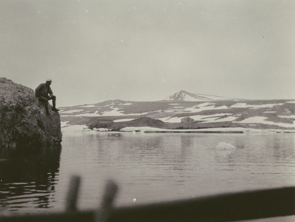 Fotografi från Ahlmannexpeditionen 1931. Motiv av man som sitter på klippa bredvid vatten och snötäckt berg.