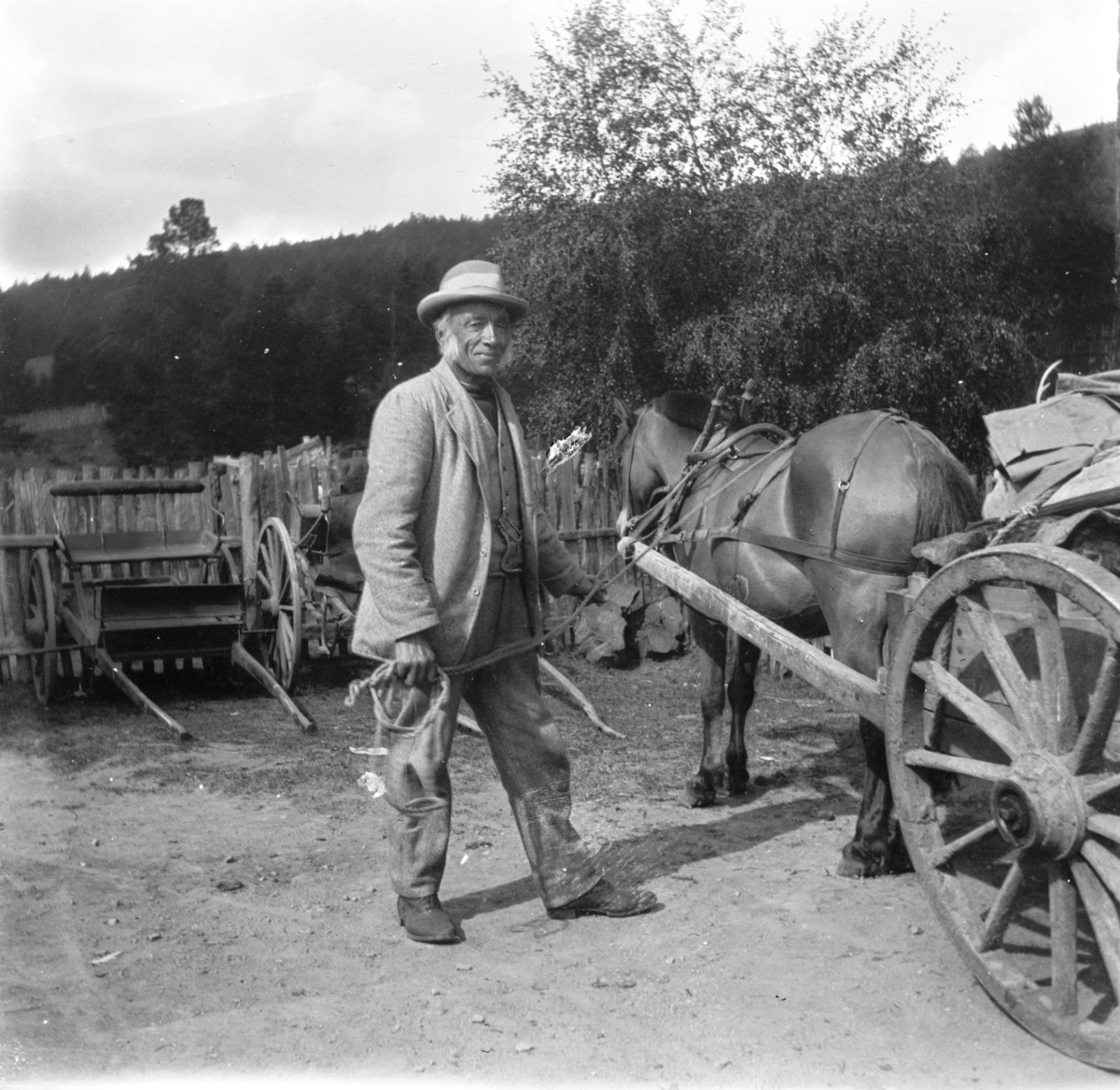Jens Tronhus i vadmelsdress og hatt står ved en hest spendt foran ei kjerre med lass