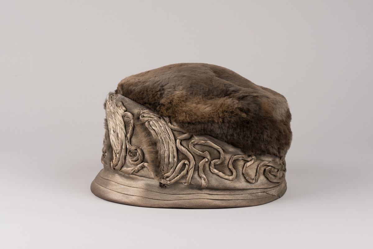 Hatt, modell med brun päls på kullen och grått tyg av metalltråd. Hatten har ett litet styvt brätte och en stärkt kant som är bredare i framkant. Dekorationer av tvinnat tyg och päls.