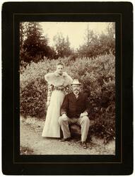 En ung kvinne står ved siden av en sittende herre i en park.