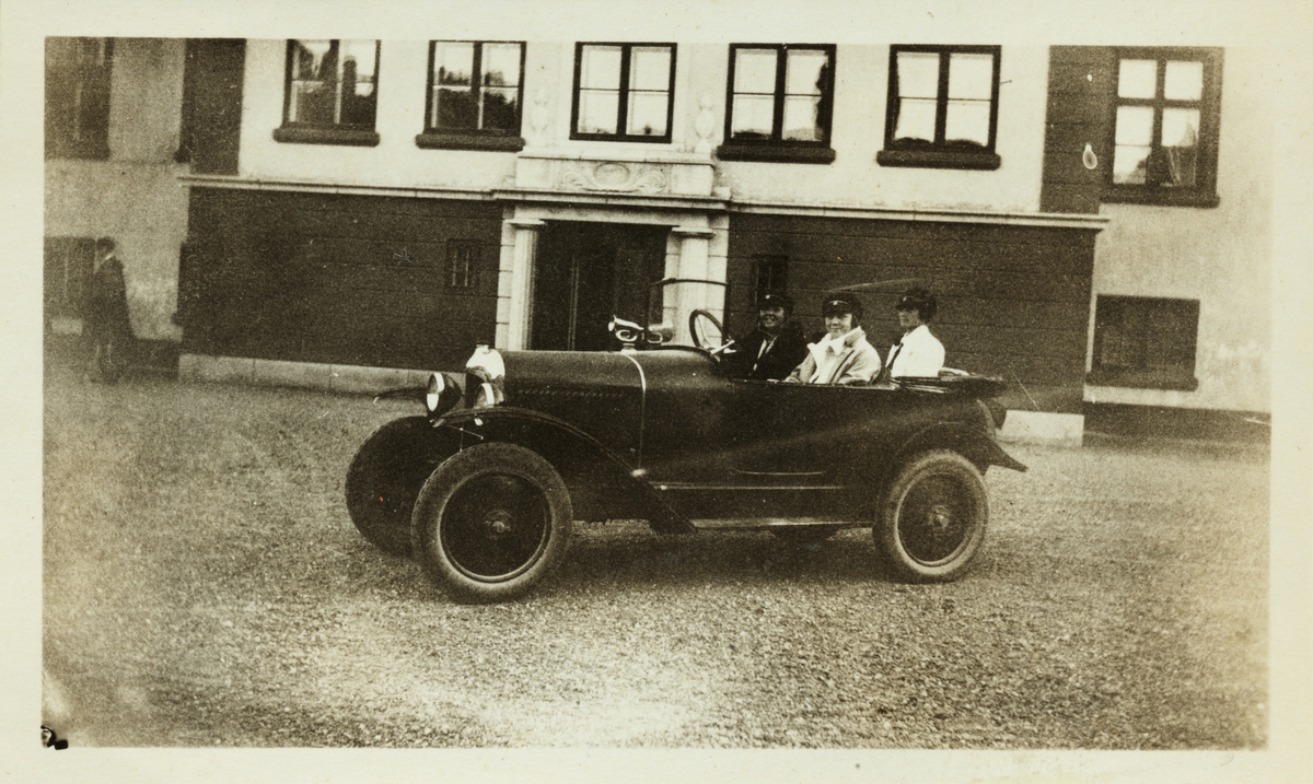 Tre ferske studenter i åpen bil på gårdsplassen til Bogstad gård. Sjåfør er Sisken Skjelderup og passasjerer er Cecilie Broch og Lucy Egeberg. Fotografert 2. september 1926.