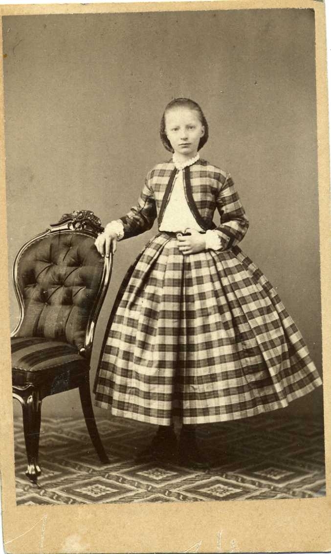 En flicka i rutig klänning och livstycke står vid en stoppad stol.