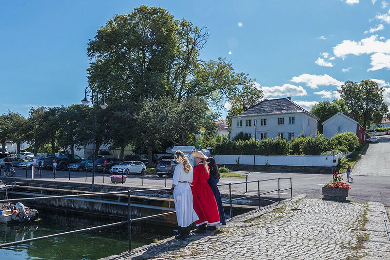 Vi ser den berømte broen i Åsgårdstrand, fra maleriet til Munch Pikene på broen. I bildet står tre damer med kjoler og hatter. Det er sommer og sol.