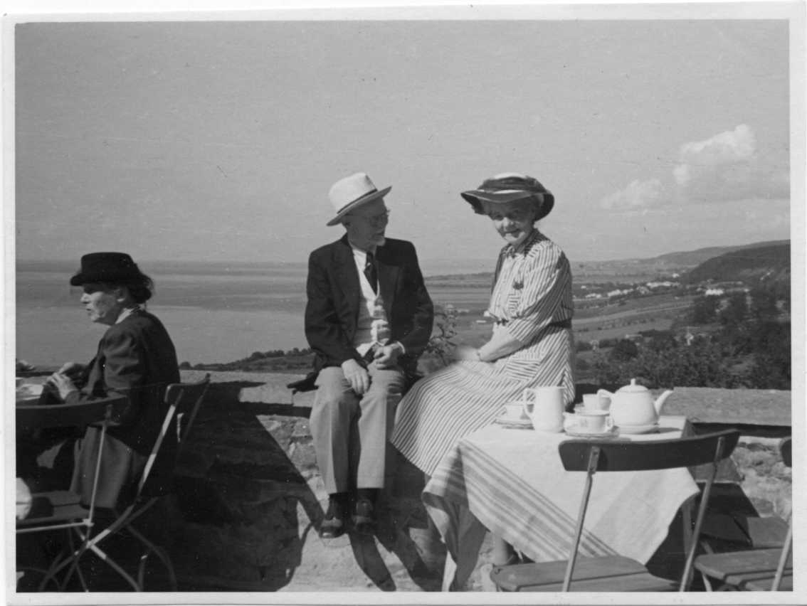 En äldre man och kvinna sitter i solsken på muren till en terass med servering. Nedanför dem står ett bord med kaffekanna och koppar. Utsikt norrut mot Gränna.