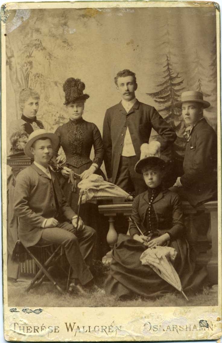 Ett sällskap med tre unga kvinnor och män i en ateljé, klädda för promenad med hattar, parasoll och spatserkäpp.