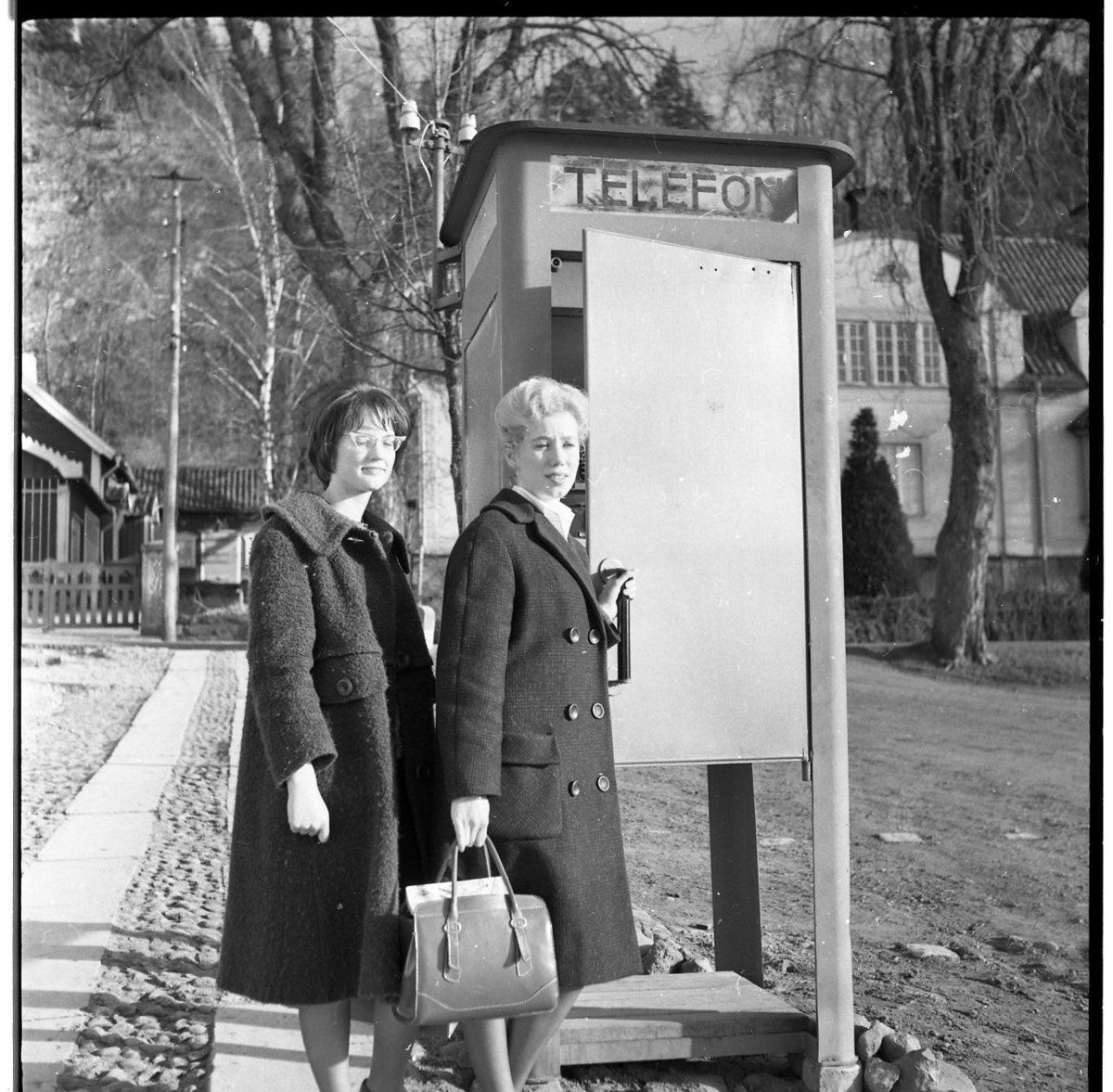 Två kvinnor står utanför en telefonkiosk. I bakgrunden skymtar Grennaskolan. Till höger står Anita Sandström.