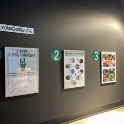 Bilde fra utstillinga "Menneskeskapt" viser tre plakater på en vegg med nummer på veggen. Vi ser ikke hva som er på plakatene.